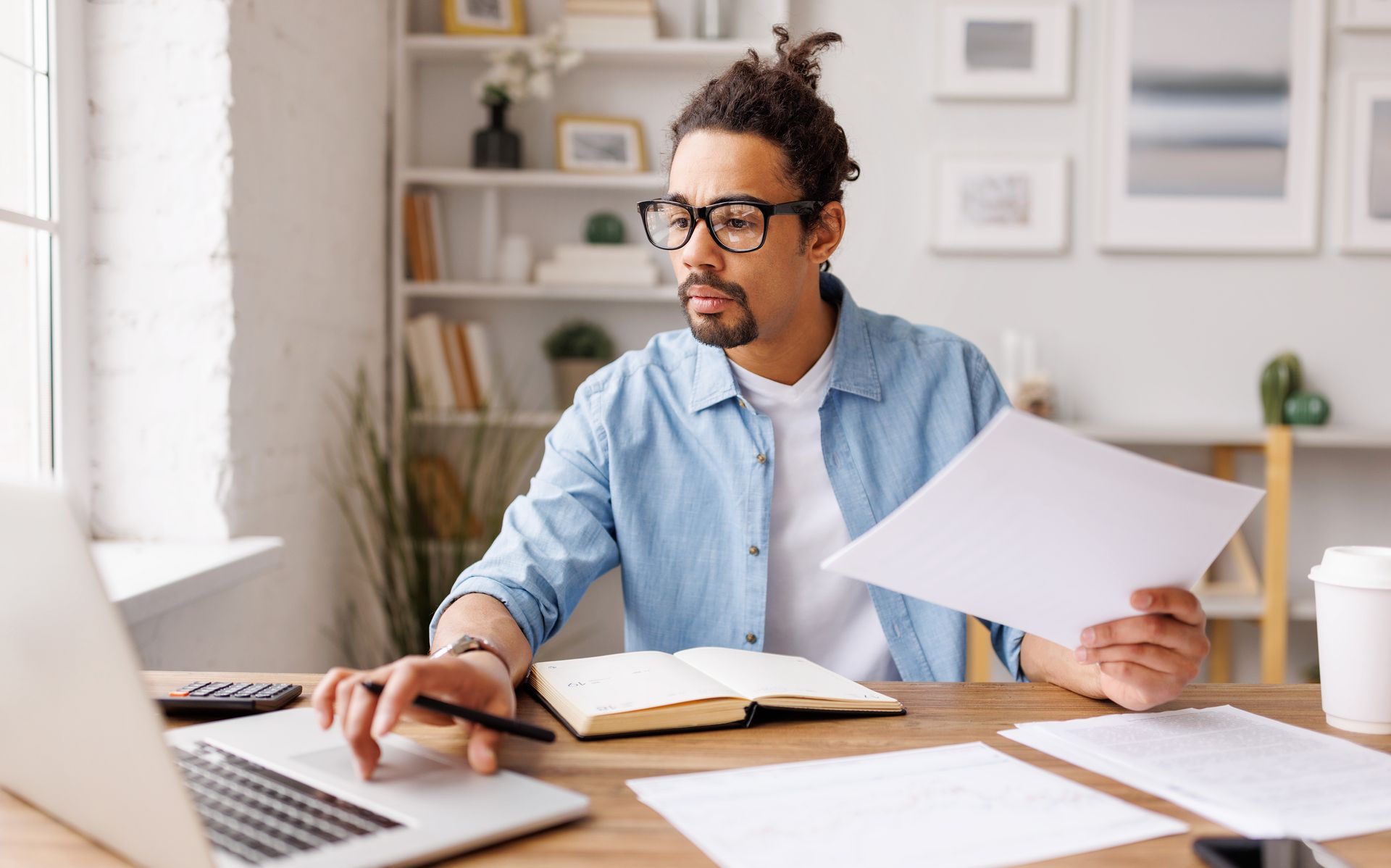 Man with glasses working on laptop, reviewing documents at a desk in a home office.