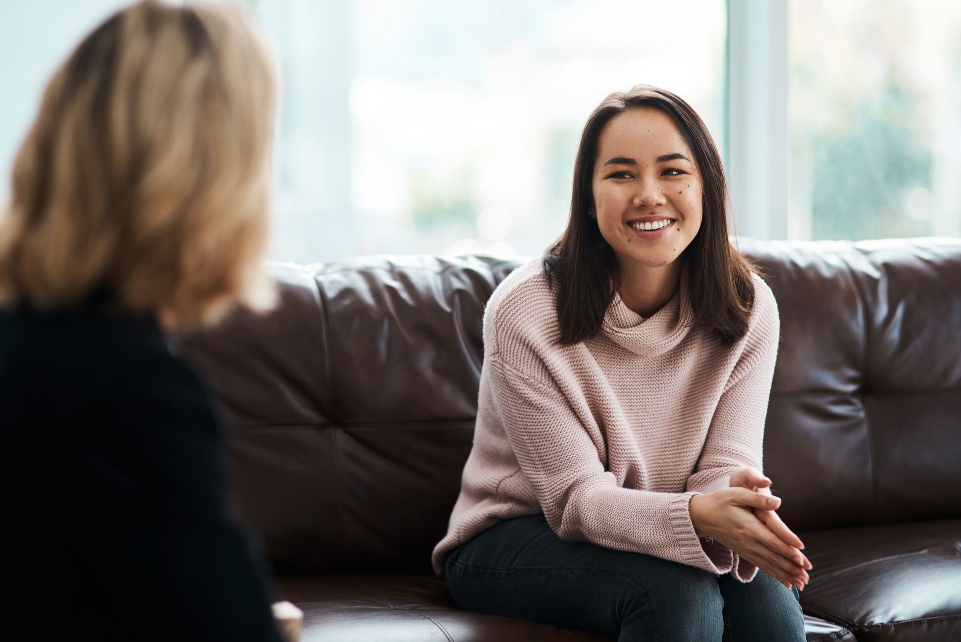 Woman smiles while sitting on a couch, facing away from a person with a black jacket.