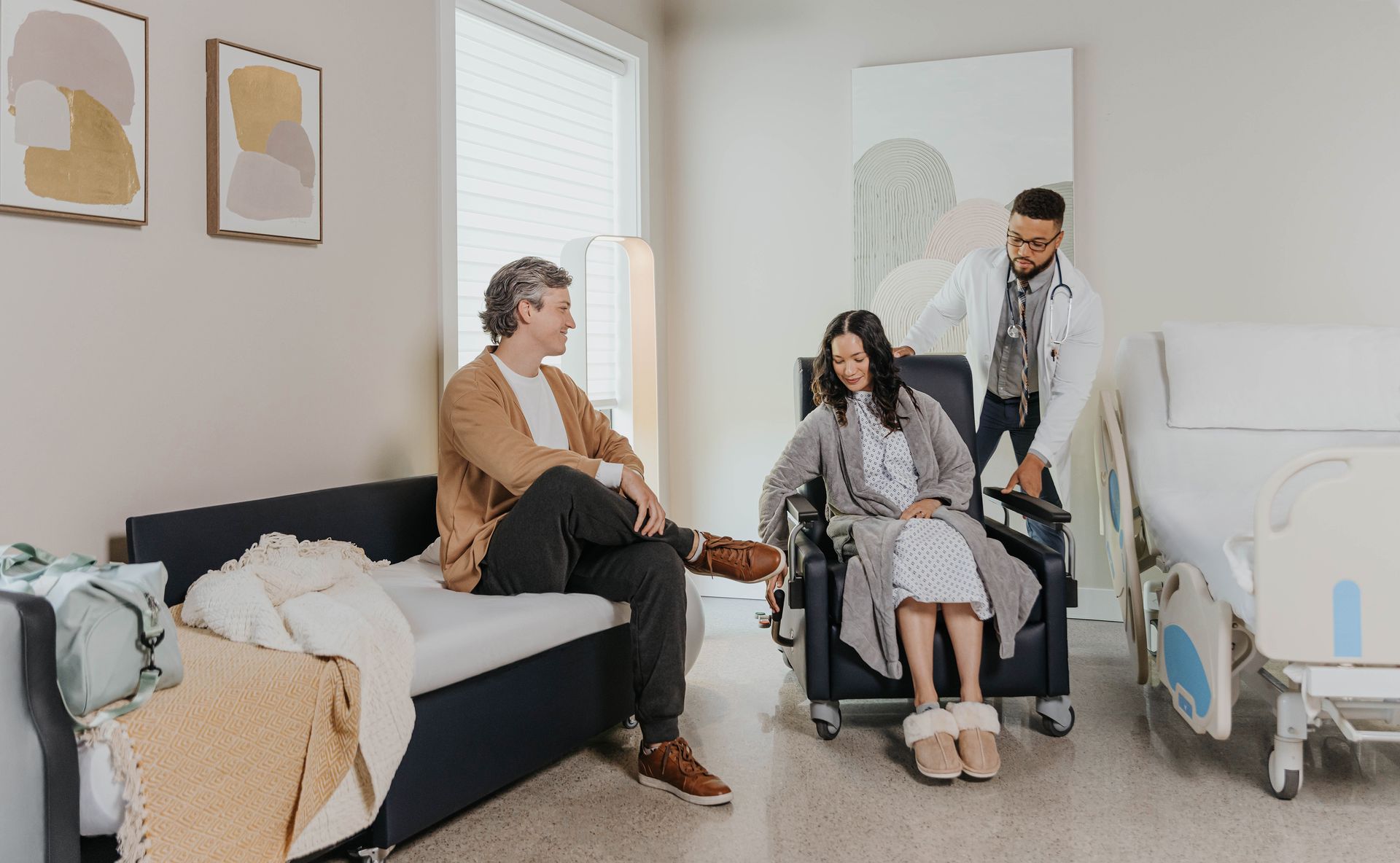 A doctor assisting a woman in a wheelchair while a man sits on a couch in a hospital room.