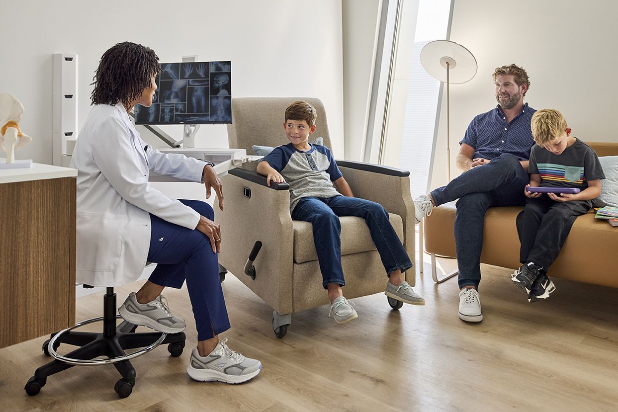 Doctor talking to a boy in a chair. Father and another boy on a couch in a medical office.