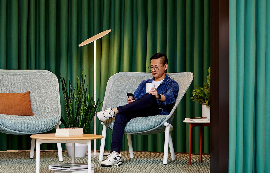 A person with glasses sits in a modern chair, looking at a phone, in a room with green curtains.