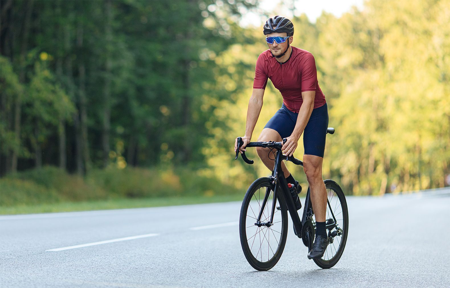 Cyclist in red shirt and blue shorts rides a black bike on a road with green trees in the background.