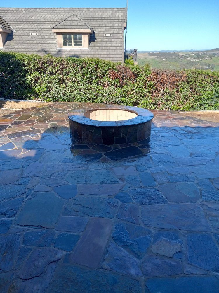 Circular fire pit on a blue-gray stone patio with a house and hillside in the background.