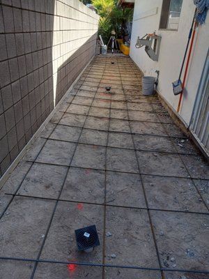 Construction site: narrow walkway with rebar grid laid on sand, bordered by concrete block wall and white building.
