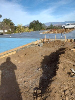Construction site with dirt, concrete slab, and wooden framing. A shadow of a person is in the foreground.