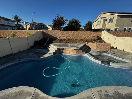 Swimming pool in backyard with retaining wall, stairs, and houses in the background under a blue sky.