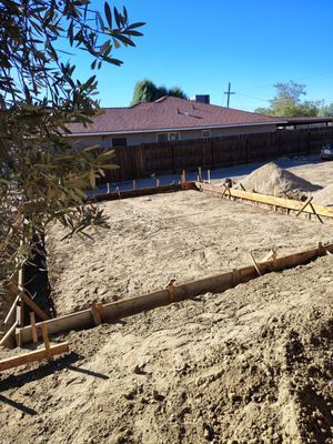 Construction site: wooden forms outline a rectangular foundation on a dirt lot, brown fence, blue sky.