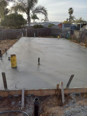 Freshly poured concrete foundation in a backyard, surrounded by wooden framing and a fence.