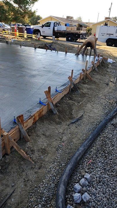 Construction worker smoothing wet concrete, wooden frame supports. Trucks in background.