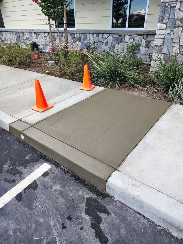 New concrete sidewalk ramp with orange safety cones; building in background.