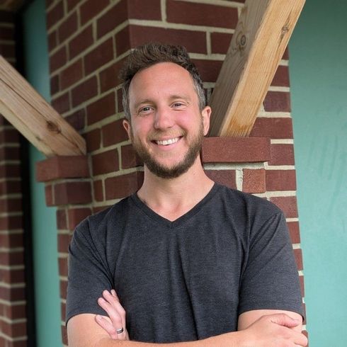A person wearing a beige sweater and beanie kisses a Boston Terrier in a matching yellow knit sweater against orange.