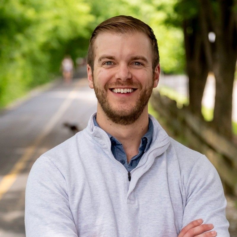A person with dark, voluminous curly hair smiles with eyes closed, wearing a patterned top against a wood-paneled wall.
