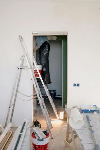 A person painting a doorway, standing on a ladder inside the door frame. Various painting supplies and tools are visible.