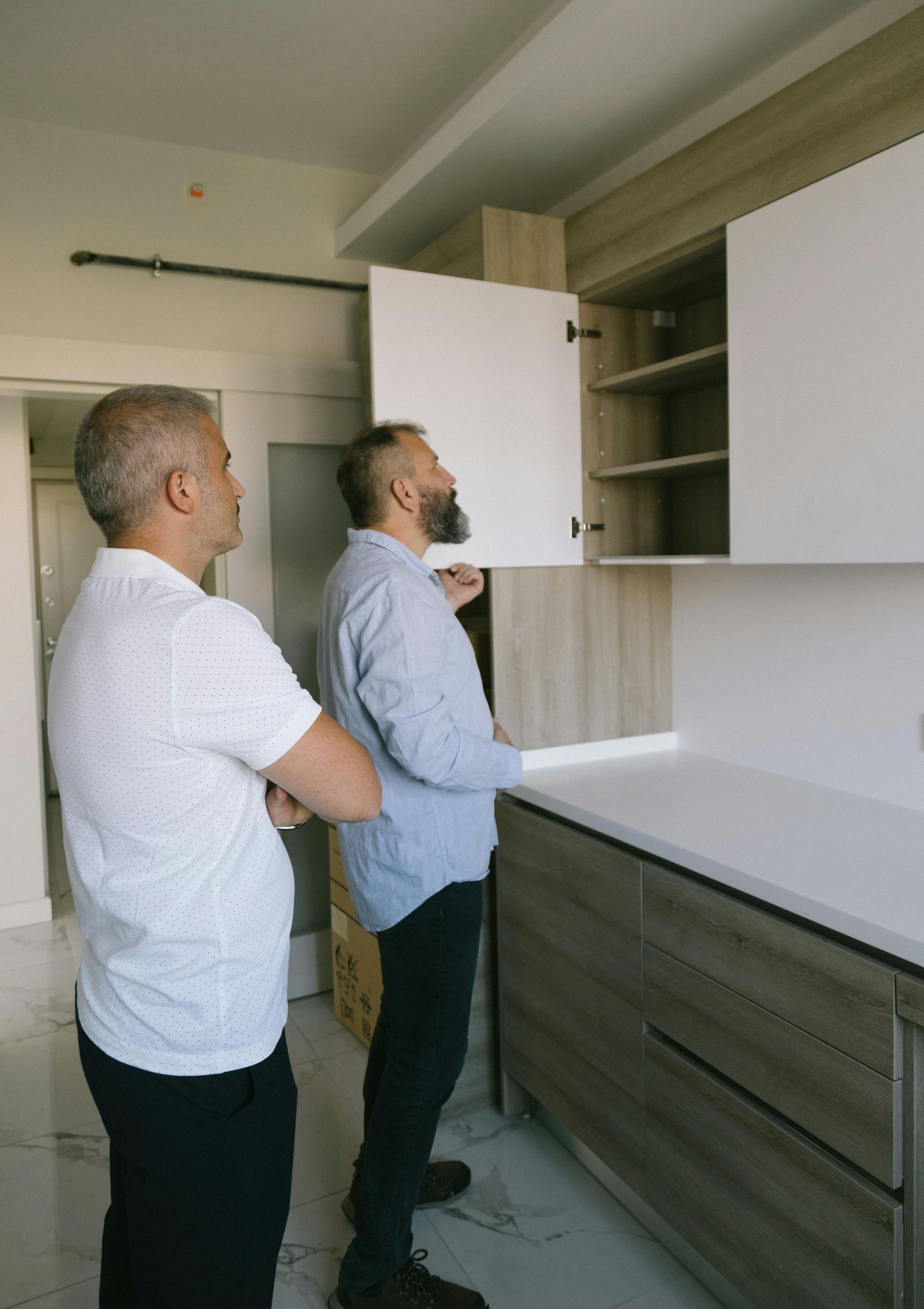 Two men inspecting a new kitchen. One opens a cabinet, examining the shelves. White and wood-tone cabinets.