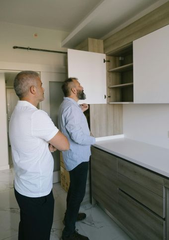 Two men inspecting a new kitchen. One opens a cabinet, examining the shelves. White and wood-tone cabinets.