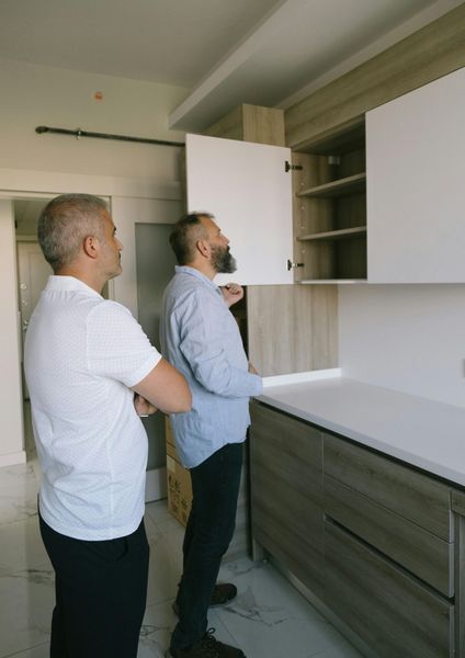 Two men inspecting a new kitchen. One opens a cabinet, examining the shelves. White and wood-tone cabinets.