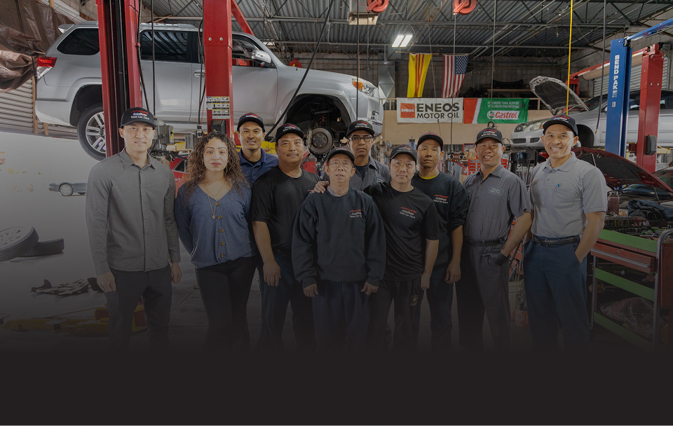 A group of nine mechanics in uniform poses for a photo inside an automotive repair shop with a car on a lift. | Japanese Auto Pros