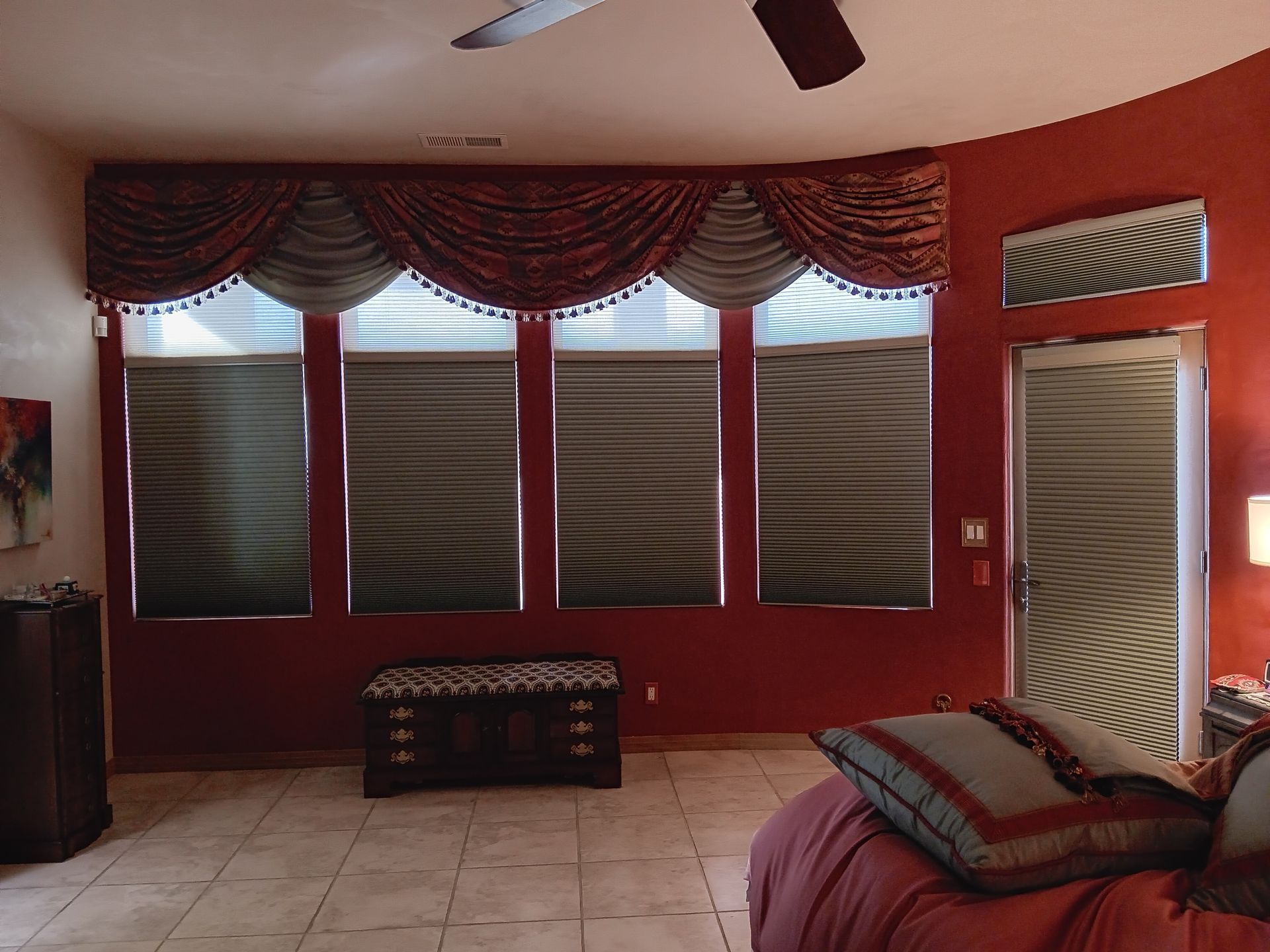 A bedroom with a red wall, windows with shades, and a decorative valance. A bed with a pillow is in the foreground.