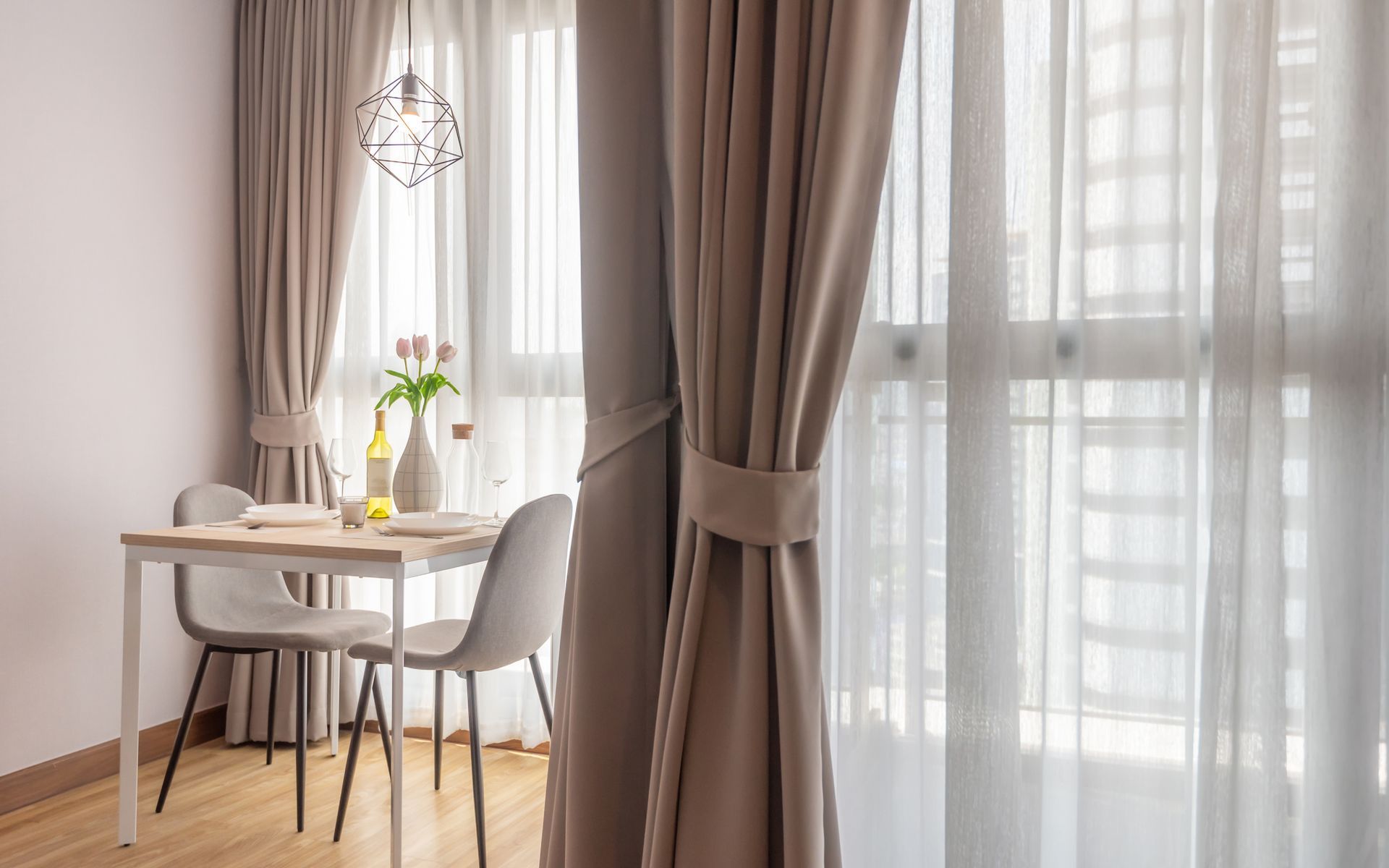 Dining area with a small table, two chairs, and sheer and solid beige curtains. Sunlight streams in.