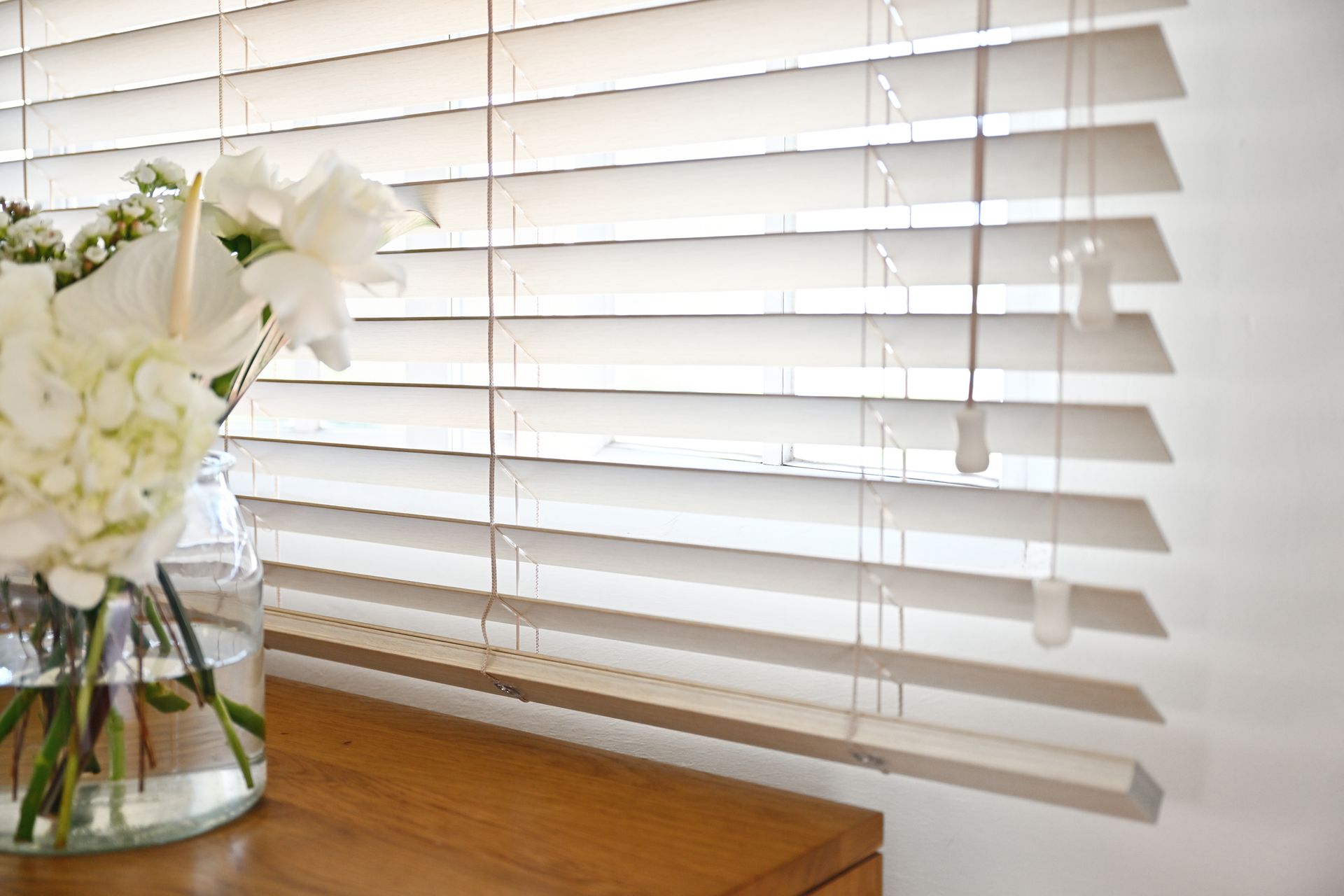 White blinds with white flowers in a glass vase on a wooden surface next to a wall.