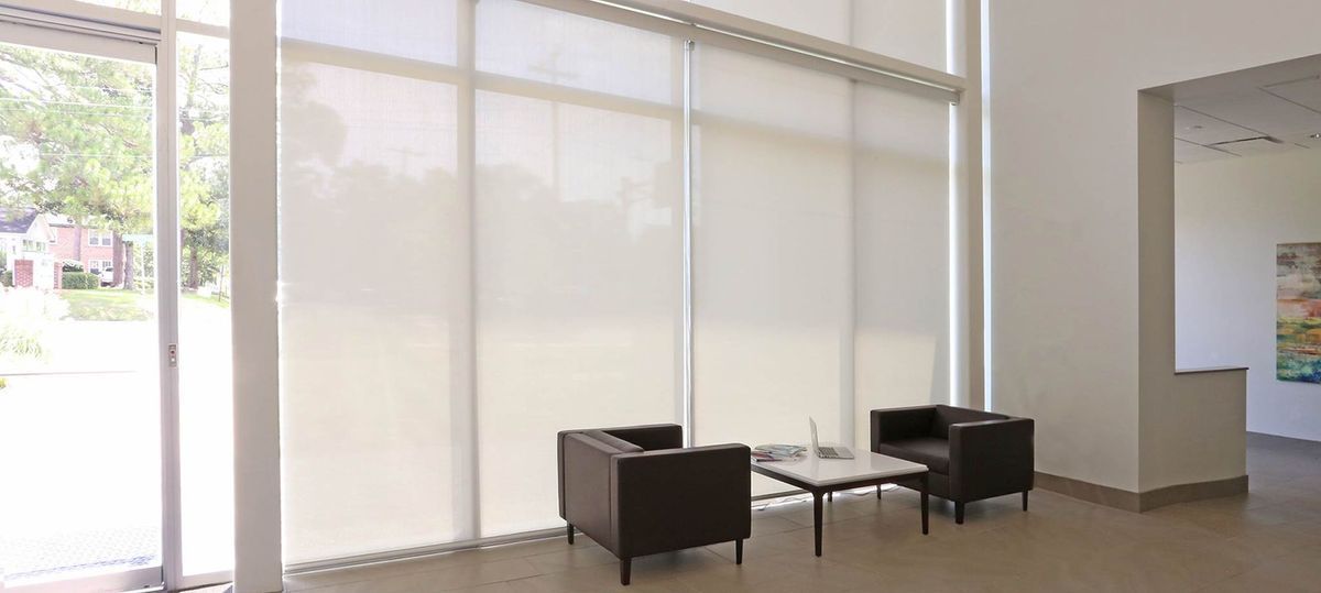 Two dark chairs and a small table in front of large windows with shades. The setting is a modern, well-lit space.