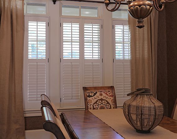 Dining room with shuttered windows, tan curtains, wooden table with centerpiece, and patterned chair.