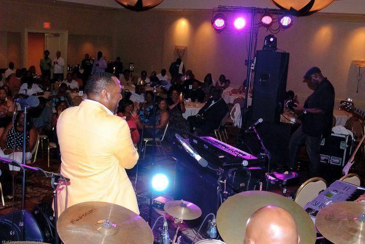 Musicians performing onstage at a crowded indoor event, with purple lights and audience seated at tables
