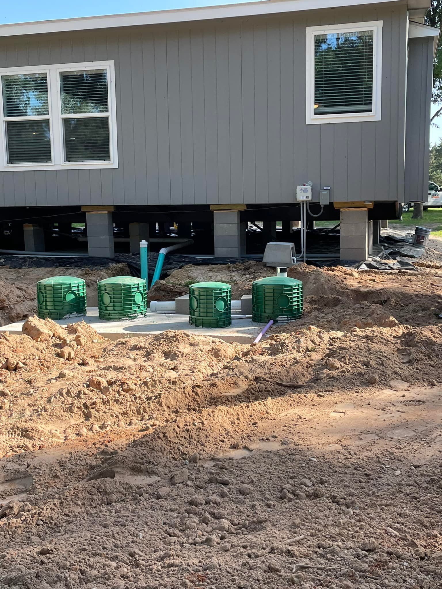 Gray house on cinder blocks with green septic system containers in a dirt yard.