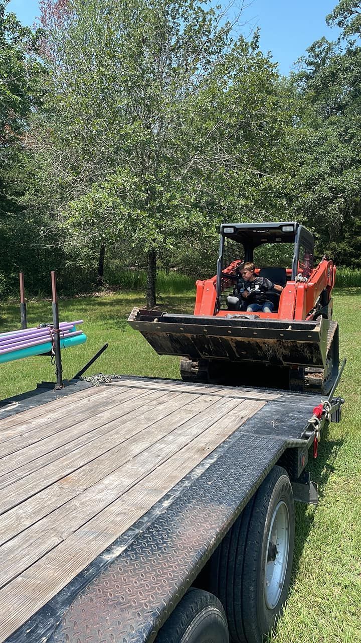 Orange skid steer loader on a trailer in a grassy area with trees.