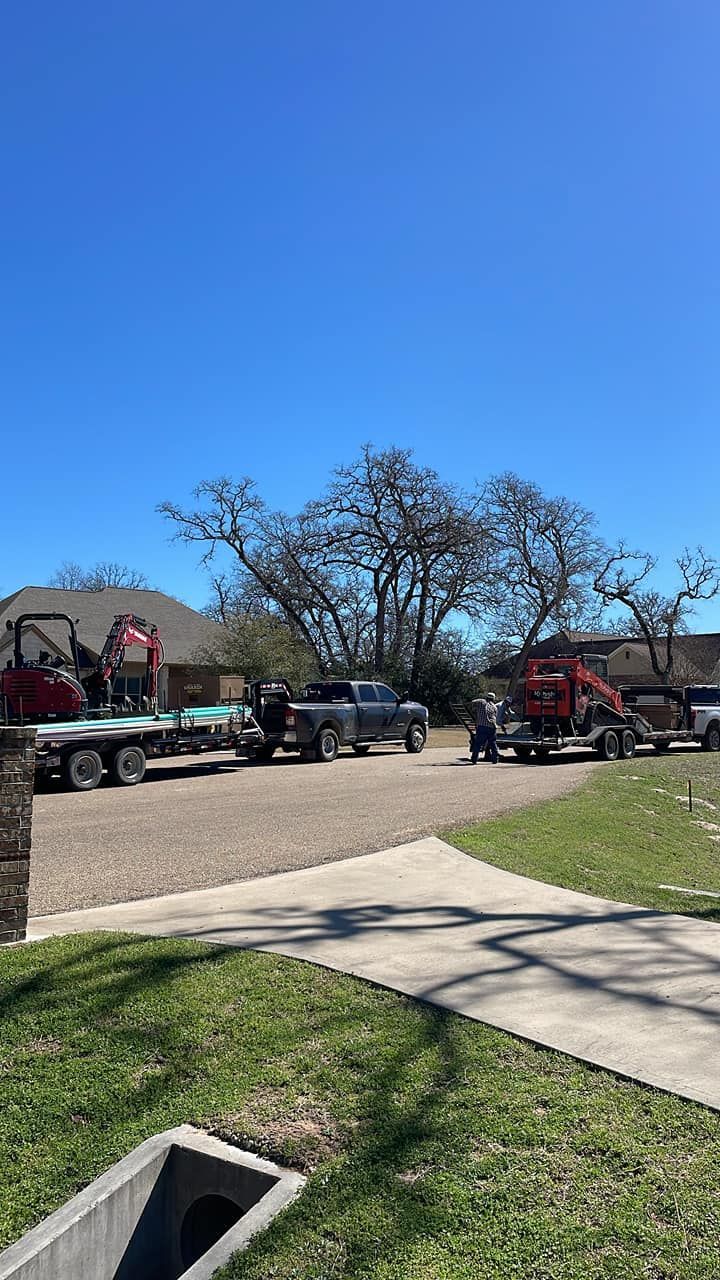 Trucks and trailers parked in front of a house on a sunny day; construction equipment visible.