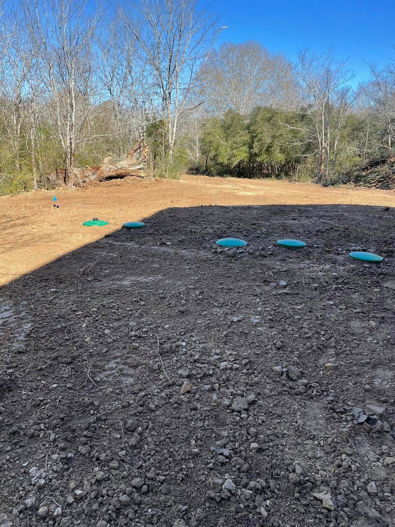 A cleared dirt lot with several blue irrigation valve covers, trees in background, under a bright blue sky.
