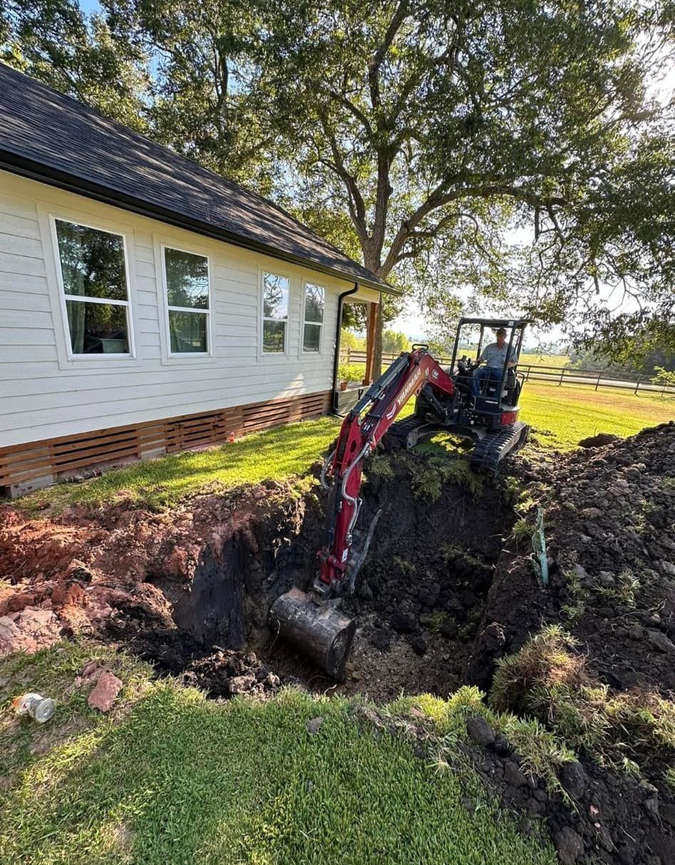 A red tractor excavates a trench beside a white house with windows and brick foundation.