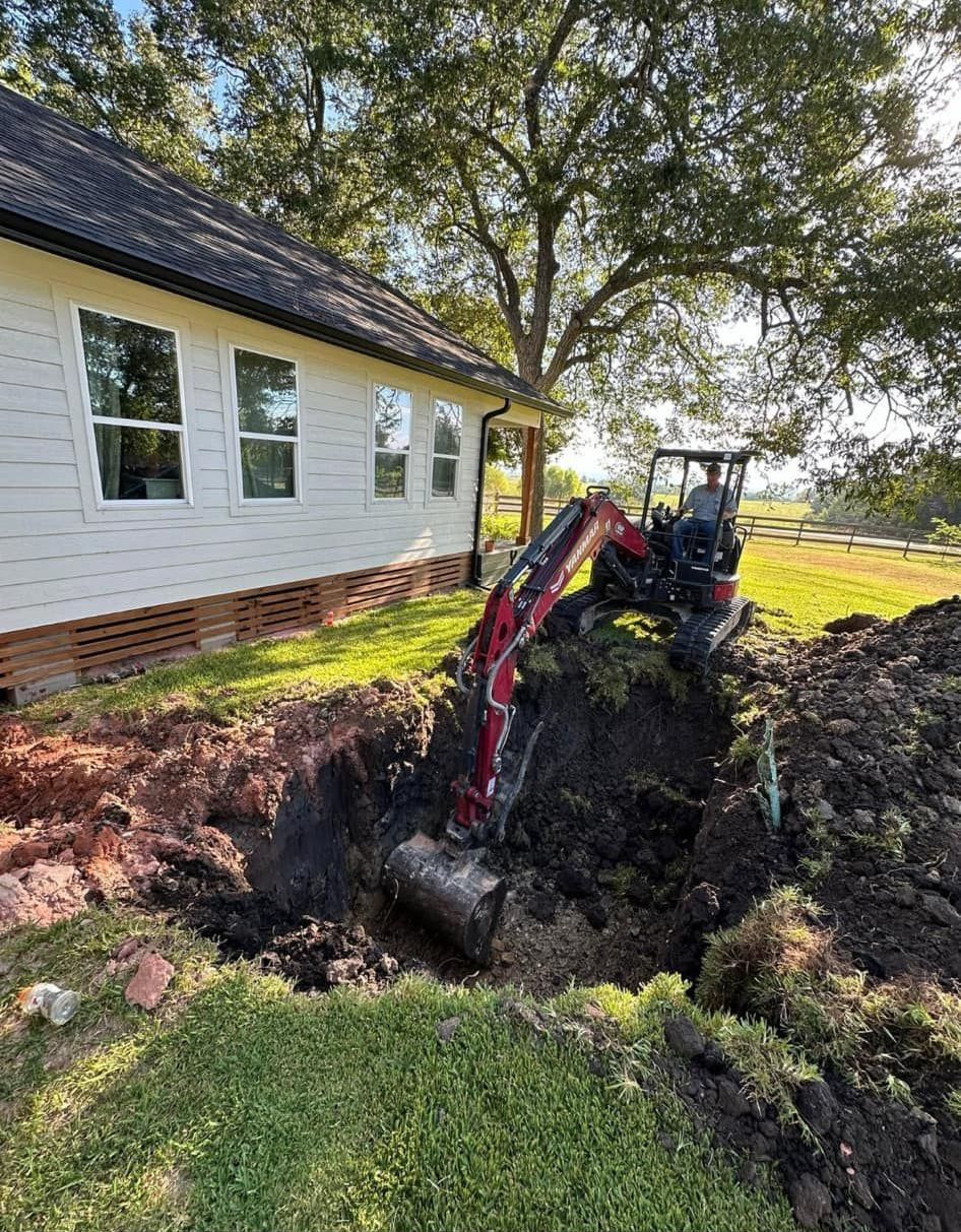 An excavator digs a trench near a white house with windows. Green grass surrounds the area.