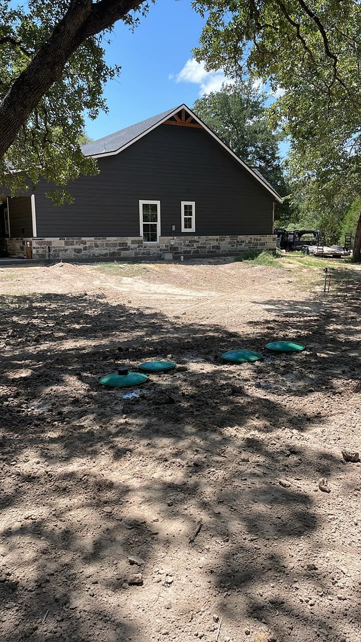 Dark gray house with visible septic lids in a yard. Sunny day.