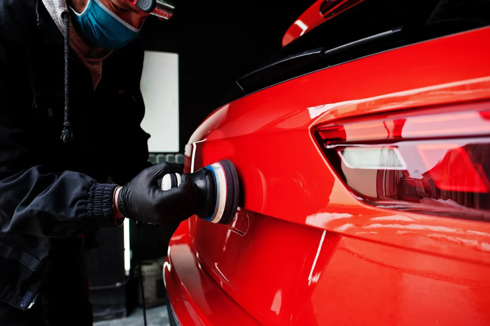 Person in black gloves polishing a red car's rear with a buffing machine in a garage. They wear a mask.