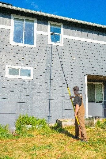 A person washes a second-story window on a gray house with a long pole, in a grassy outdoor setting. A person washes a second-story window on a gray house with a long pole, in a grassy outdoor setting.