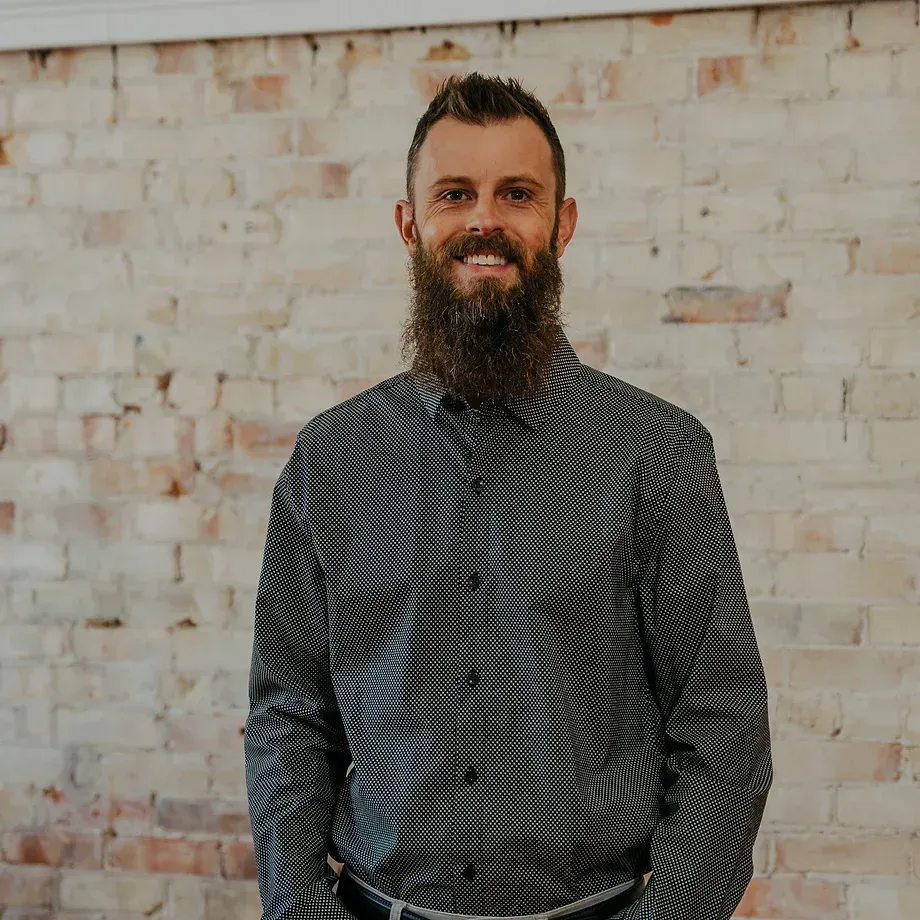 A man with a beard is standing in front of a brick wall.
