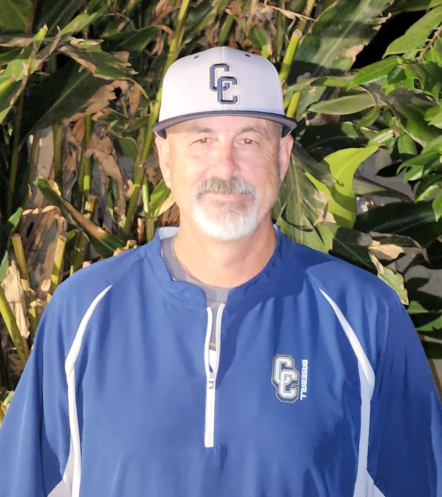 Football coach with arms crossed, looking stern on a sunny field, players in the background.