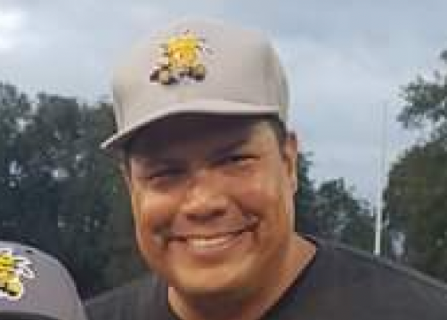 Football coach with arms crossed, standing on a football field. He wears a navy polo shirt and gray pants.