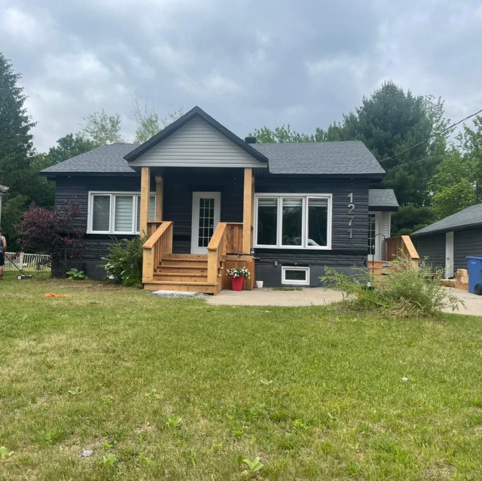 Une maison noire avec un porche et des escaliers en bois se trouve au milieu d'un champ verdoyant.