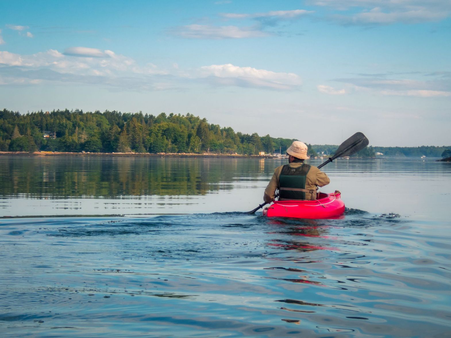 Person kayaking on a red kayak across a calm lake with forested shoreline under a blue sky