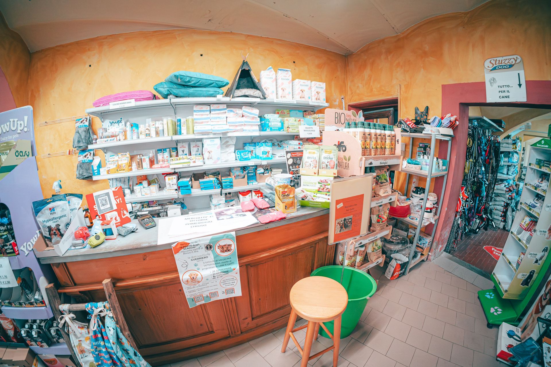 Interno di un negozio di articoli per animali, scaffali pieni di prodotti, bancone in legno, sgabello e porta aperta.