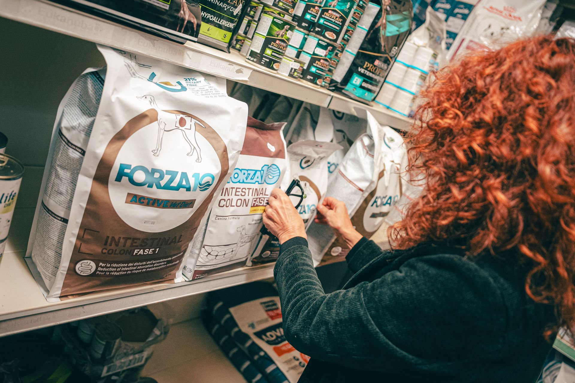 Donna con i capelli rossi e ricci che sceglie del cibo per cani da uno scaffale in un negozio di animali.