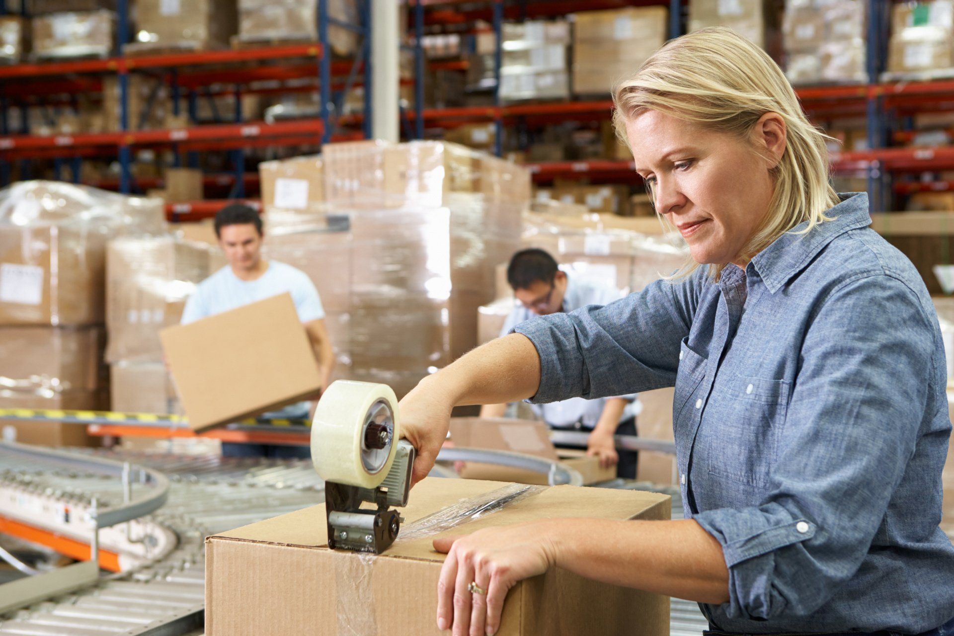 A woman is taping a cardboard box in a warehouse.