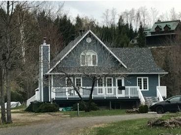 Maison grise aux boiseries blanches, avec porche et haute cheminée, située dans une zone boisée.