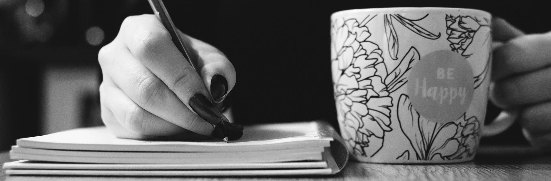 A person's hand with dark nail polish writes on a notepad with a pen; a mug with 