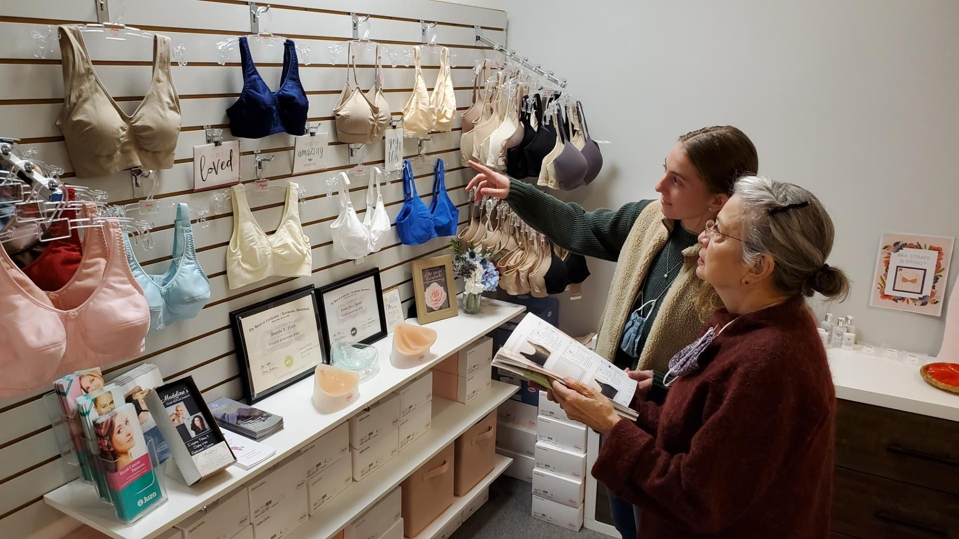 Two women in a store looking at bras displayed on a wall. One points, the other holds a catalog.