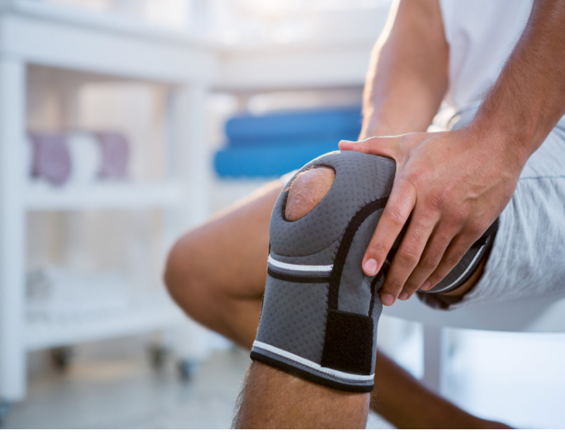 Person with knee brace holding their knee, in what appears to be a healthcare setting.