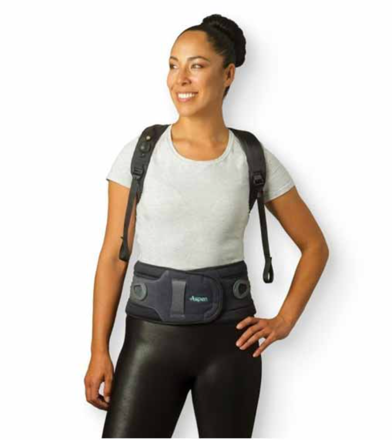 Woman smiling, wearing a back brace. She’s standing indoors, wearing a light gray shirt and black leggings.
