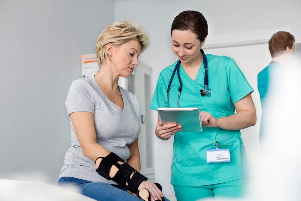 Nurse in teal scrubs shows a tablet to a woman with an arm brace in a doctor's office.
